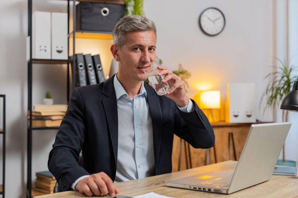 mann mit grauen haaren im anzug trinkt wasser am schreibtisch mit laptop im modernen buero, beim online business aufbauen ohne startkapital